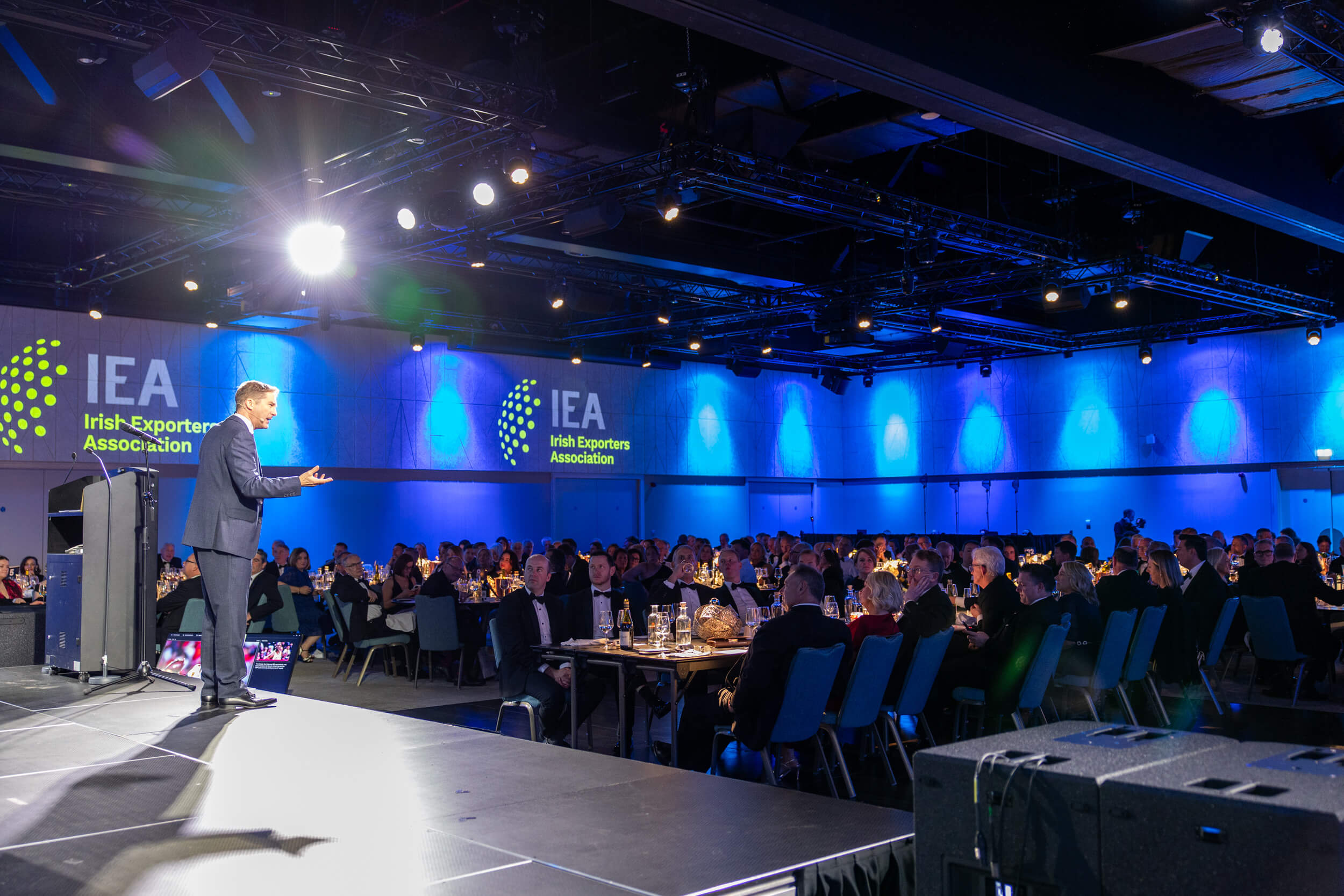 A speaker addresses an audience at a formal Irish Exporters Association gala event in a large hall with blue stage lighting.