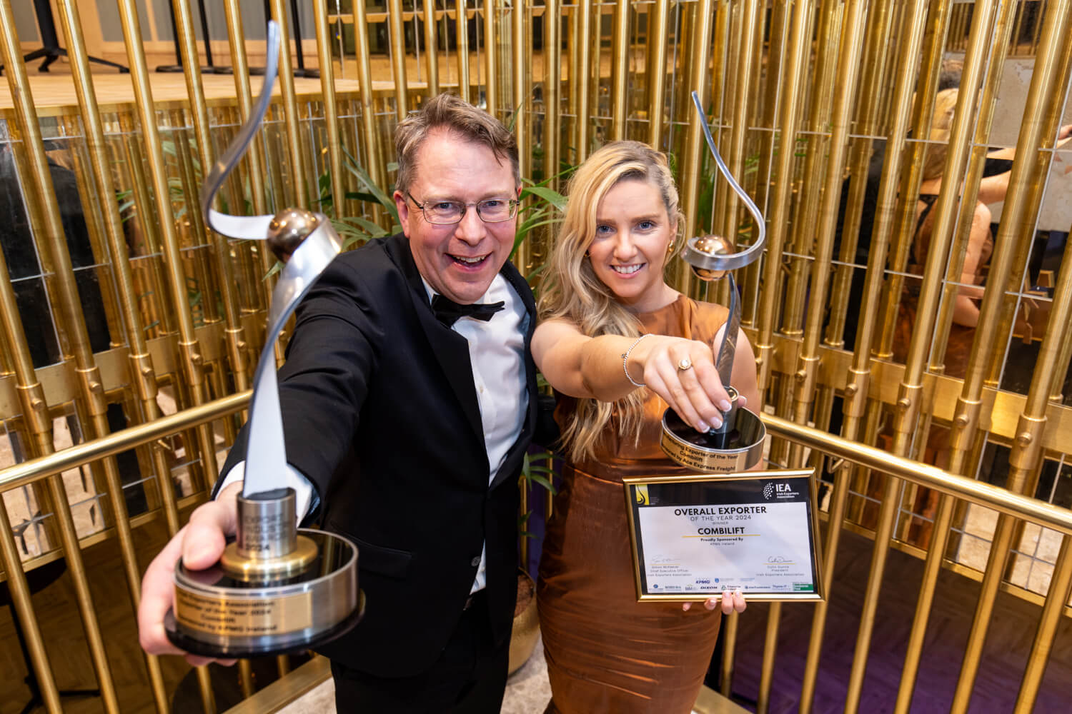 A man in a tuxedo and a woman in a copper dress holding up silver awards and a certificate for "Overall Exporter of the Year 2024."