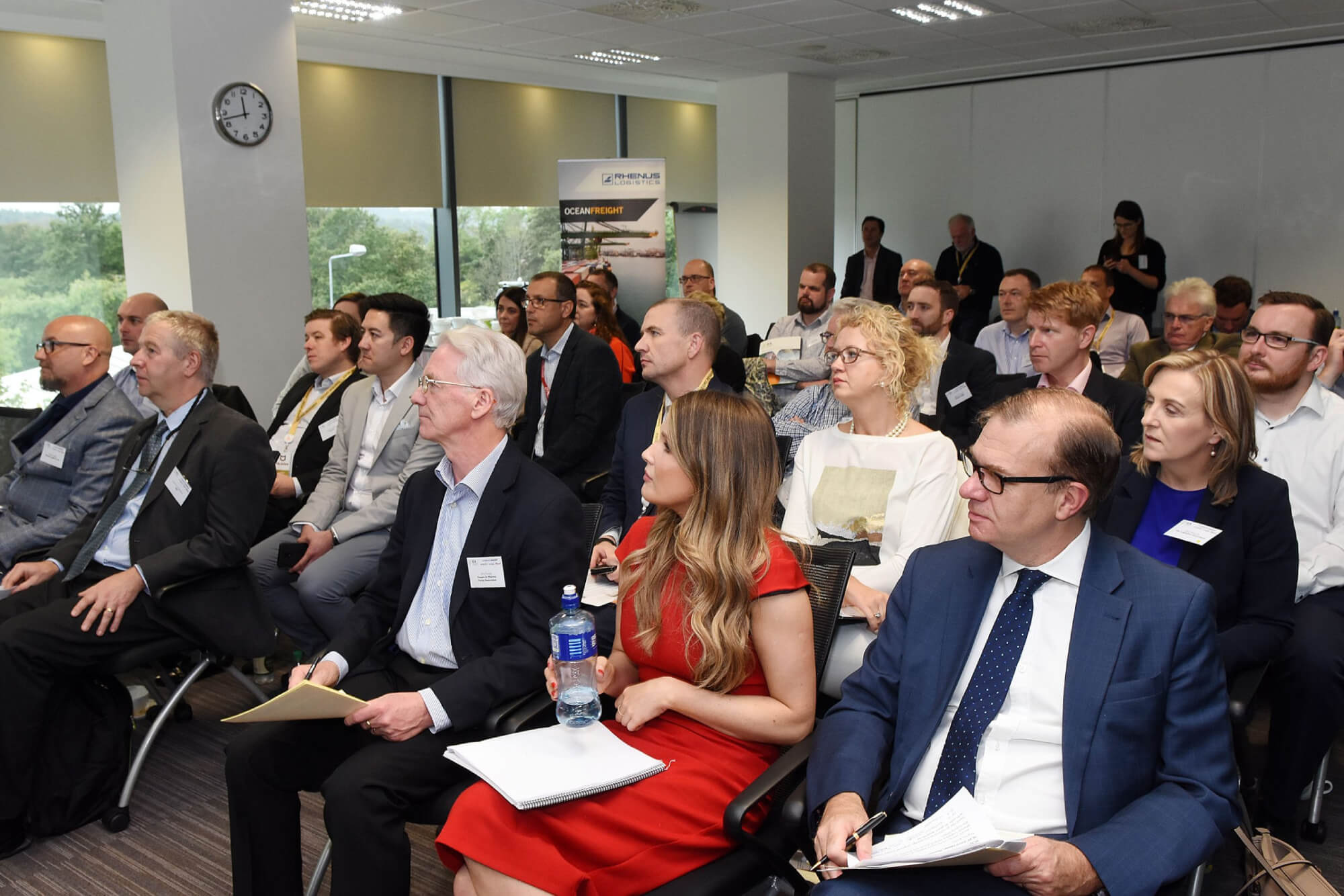 A group of professionals in business attire sit in rows of chairs, listening attentively and taking notes during a corporate presentation in a modern office setting.